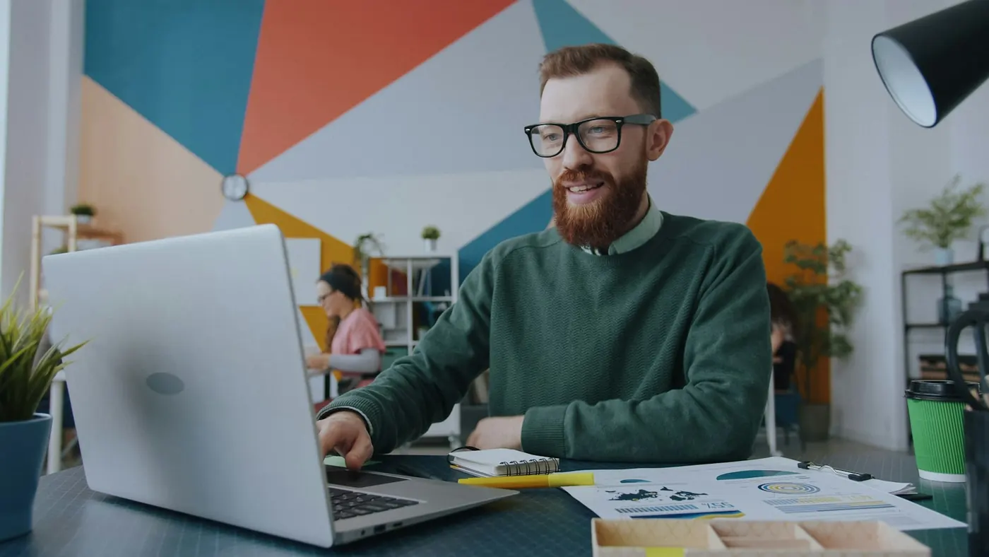 Smiling man working on a laptop in a busy creative office