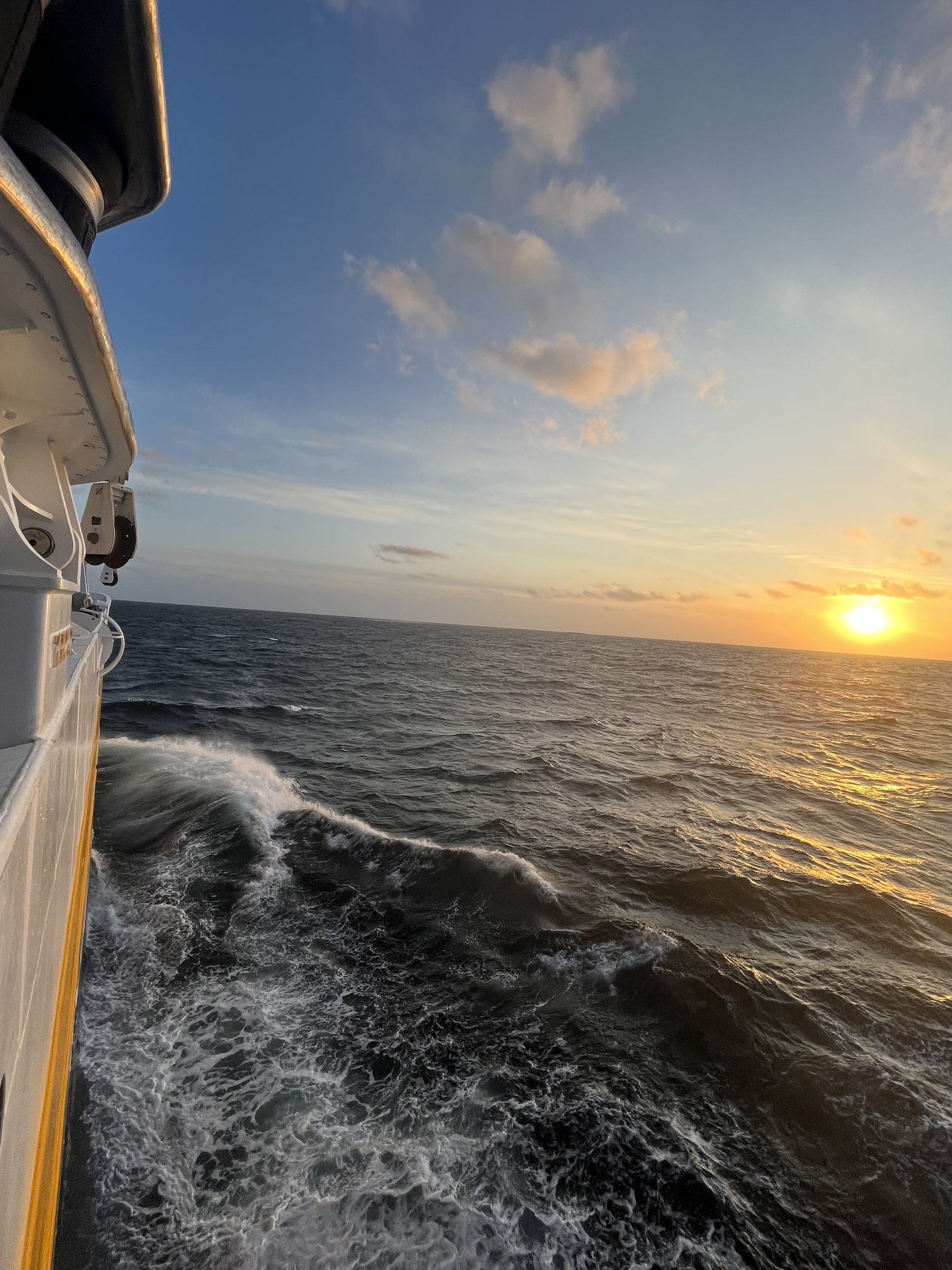 View from the side of a boat looking out at choppy ocean waves under a partly cloudy sky during sunset with the sun near the horizon reflecting golden light on the water.