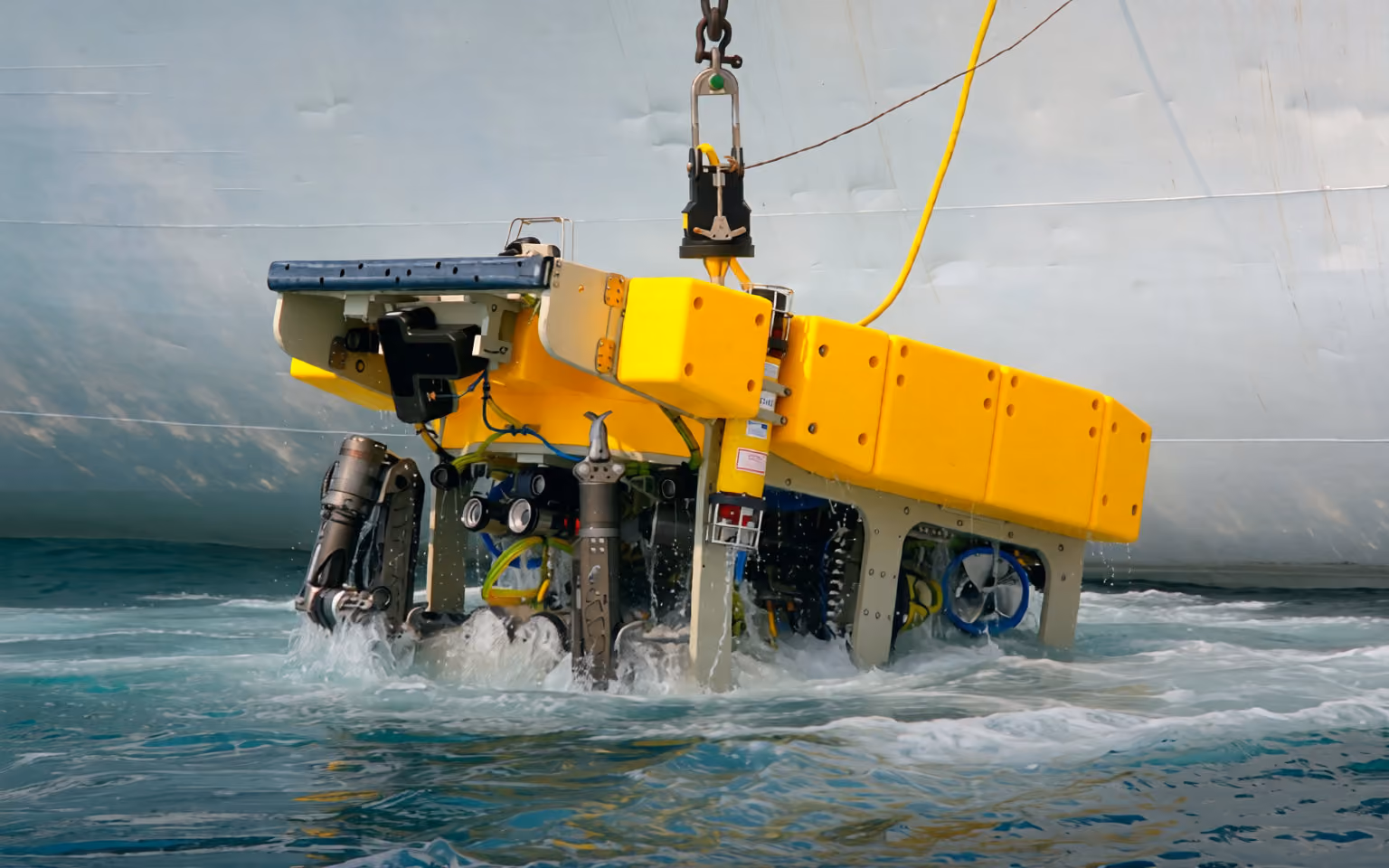 Yellow underwater remotely operated vehicle (ROV) partially submerged, being lowered into the water by a crane hook near a large ship hull.