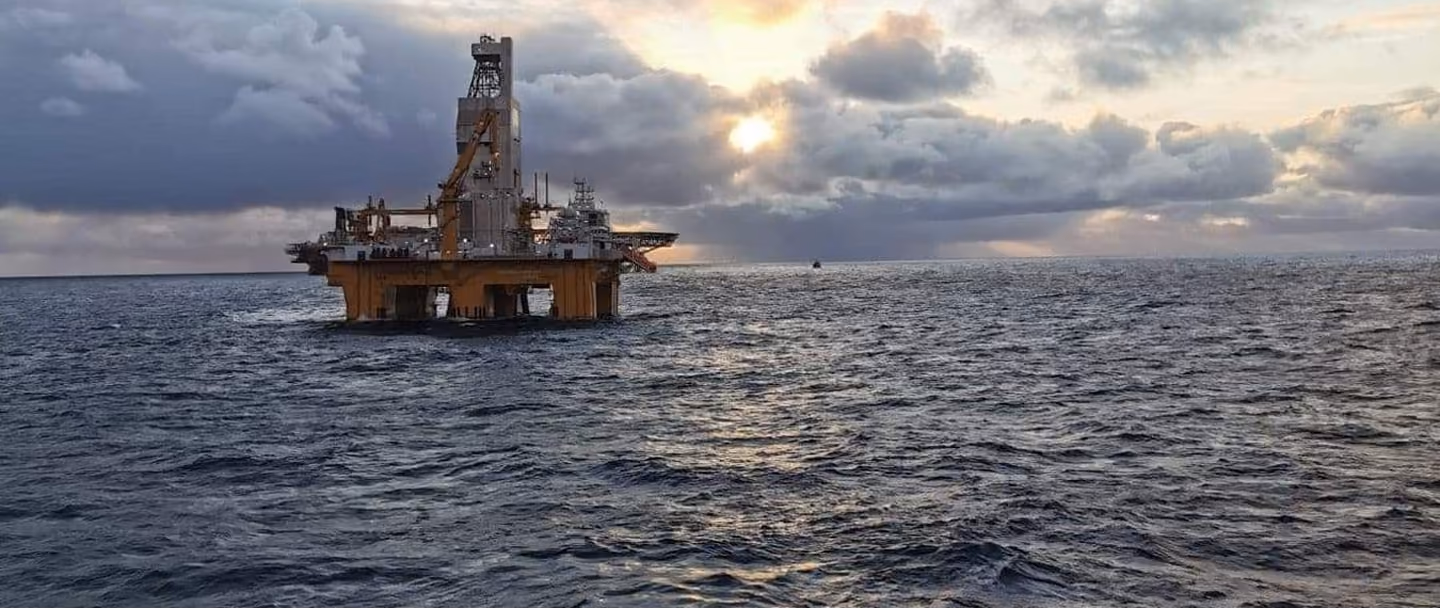 Offshore oil drilling platform in the ocean under a cloudy sky with the sun partially visible at sunset or sunrise.