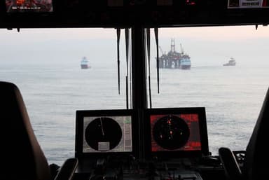 View from a ship's bridge showing two radar screens and the ocean with three ships and an offshore oil platform in the distance.
