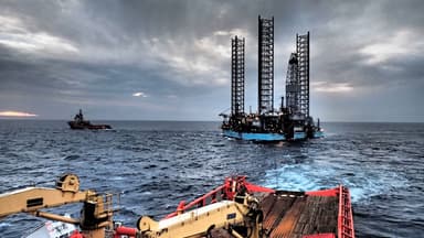 View from a ship deck looking out over the ocean at an offshore oil drilling platform with a tugboat nearby under a cloudy sky at sunset.