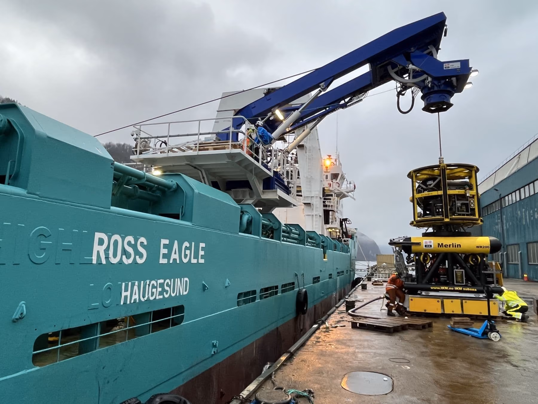 The ship Ross Eagle from Haugesund docked beside a pier with workers operating a crane lifting a yellow underwater remotely operated vehicle labeled Merlin WR200.