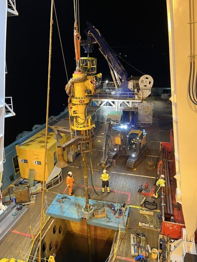 Nighttime scene on a ship's deck with workers in safety gear operating a large yellow submersible device suspended by cables above an open hatch, with heavy machinery and equipment around.