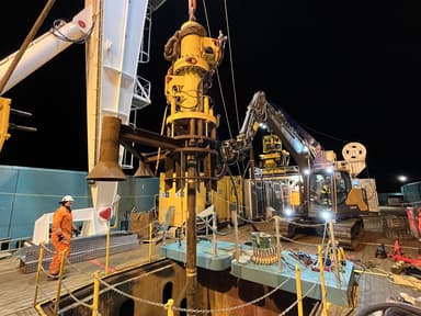 Nighttime construction site showing a large yellow drilling machine operating while a worker in an orange safety suit and white helmet observes nearby.