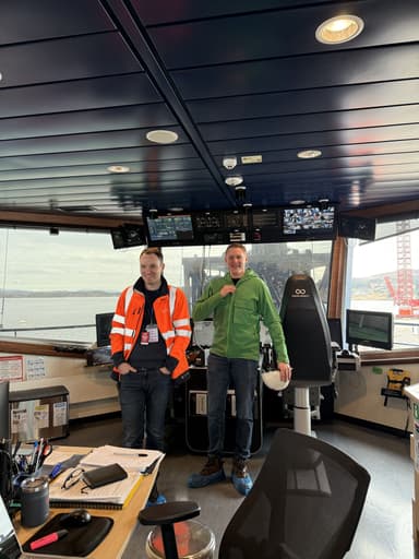 Two men standing inside a ship's control room with large windows showing a body of water and a red crane outside; one man wears a bright orange safety jacket, the other a green jacket holding a white helmet.