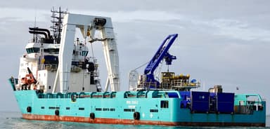 Large turquoise industrial ship equipped with cranes and machinery floating on calm water under a cloudy sky.