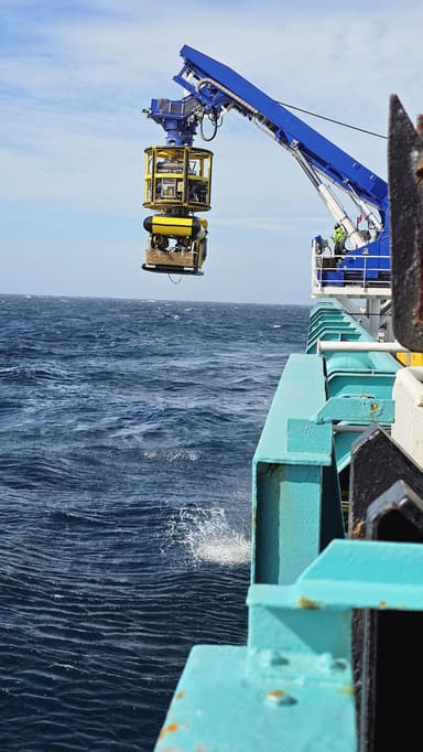 Blue crane on a ship lifting a yellow underwater exploration device above the ocean surface, with visible waves and a partly cloudy sky.