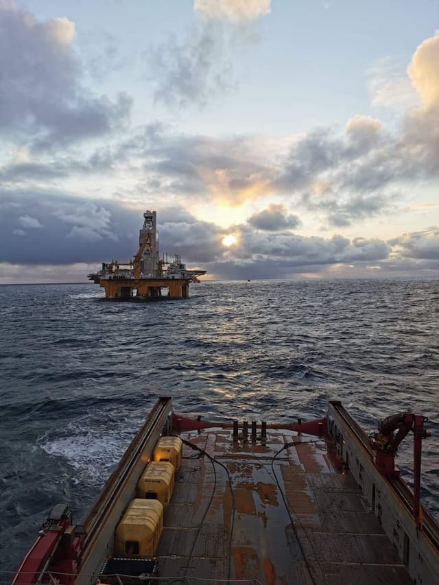 View from the deck of a vessel looking out toward an offshore oil rig at sea during a partly cloudy sunset.