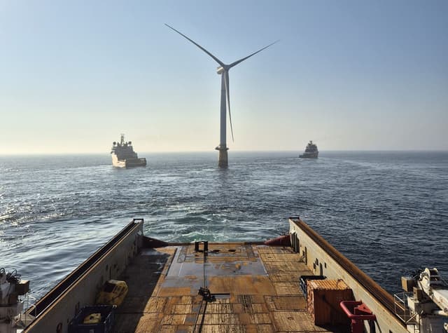 View from the deck of a ship looking out at a large offshore wind turbine in the ocean with two other ships in the distance under a clear sky.