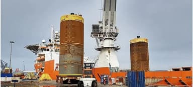 Two large cylindrical offshore platform legs being transported on a flatbed truck at a port with cranes and an orange vessel in the background under a cloudy sky.