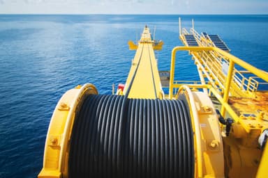 Close-up view of a large yellow industrial winch with tightly coiled black cable on a platform overlooking a calm blue sea under a clear sky.