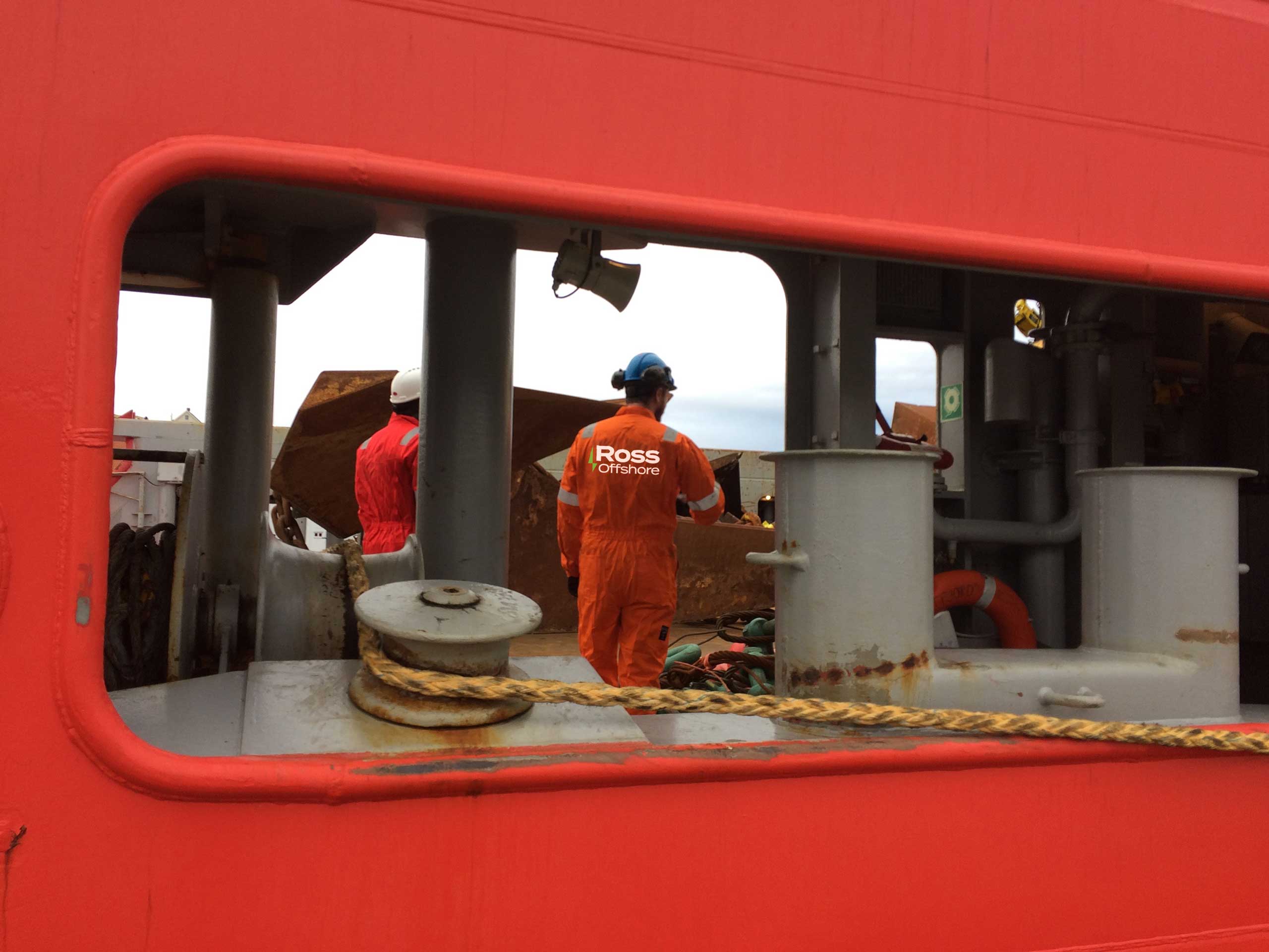 Ross Offshore crew in orange coveralls working on deck, seen through an opening in the vessel's red bulwark