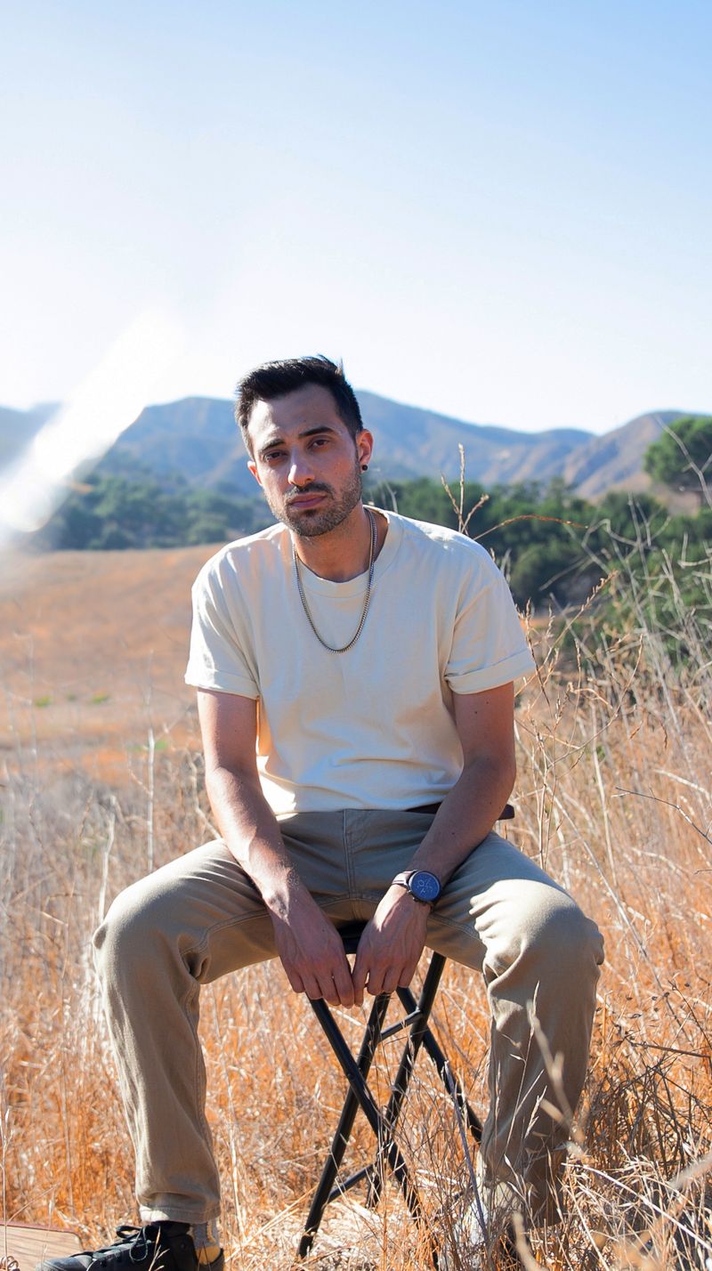 Portrait of Giosuè Greco seated on a folding stool in a field of dry grass, hills behind him