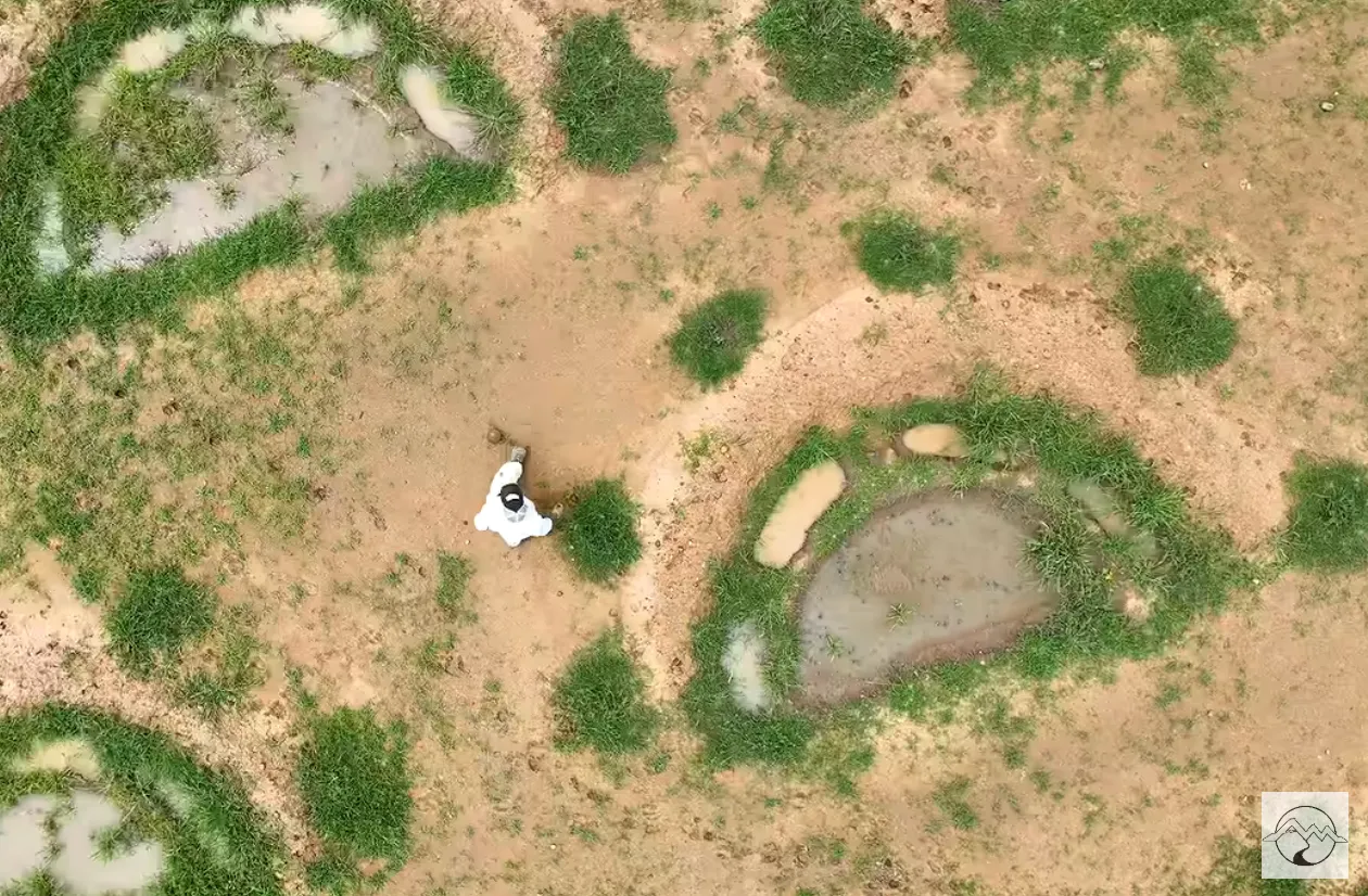 Overhead drone view of shallow catchment basins dug into dry ground, with one person standing among them for scale.