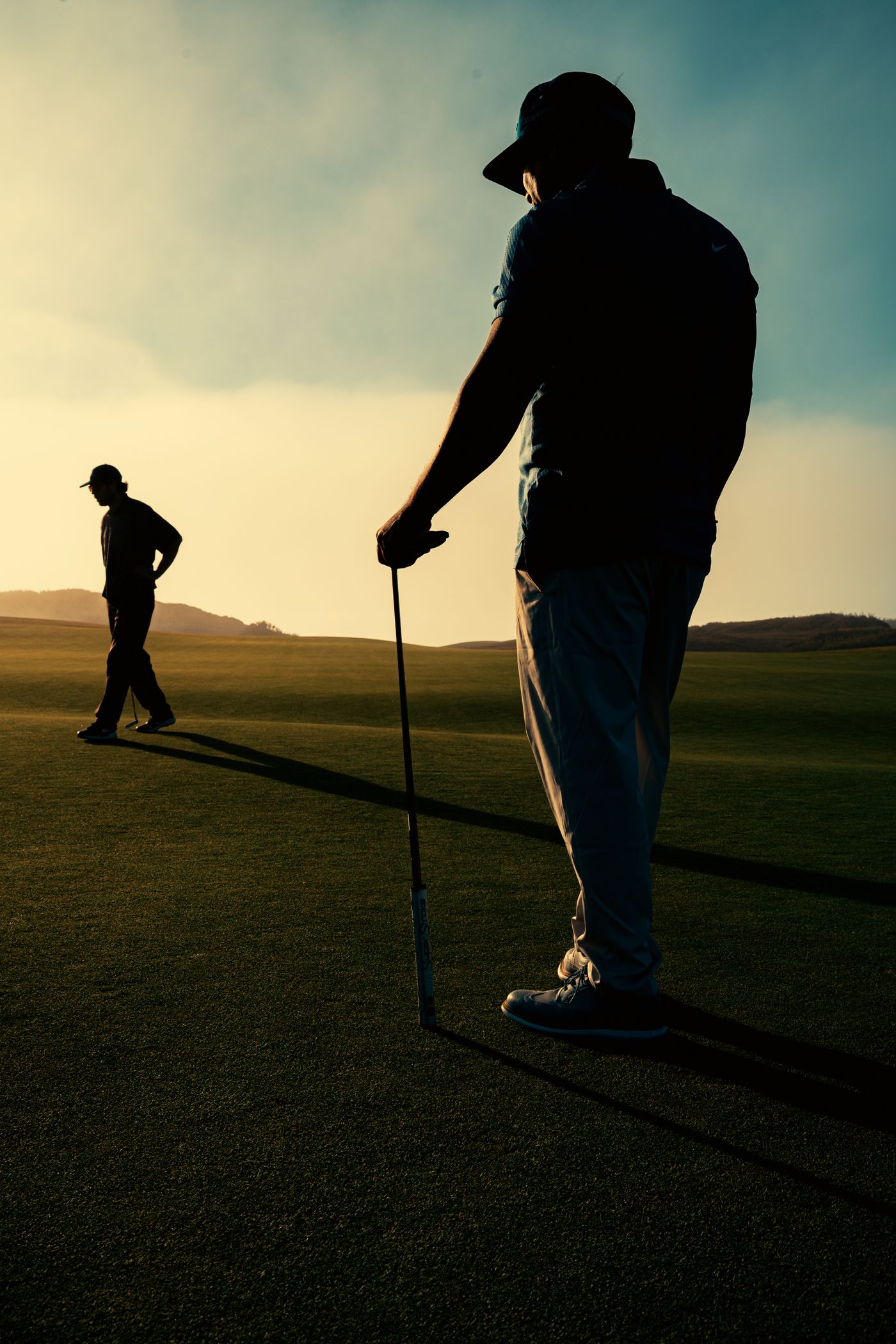 Two golfers in silhouette on a course at sunset, one leaning on a club