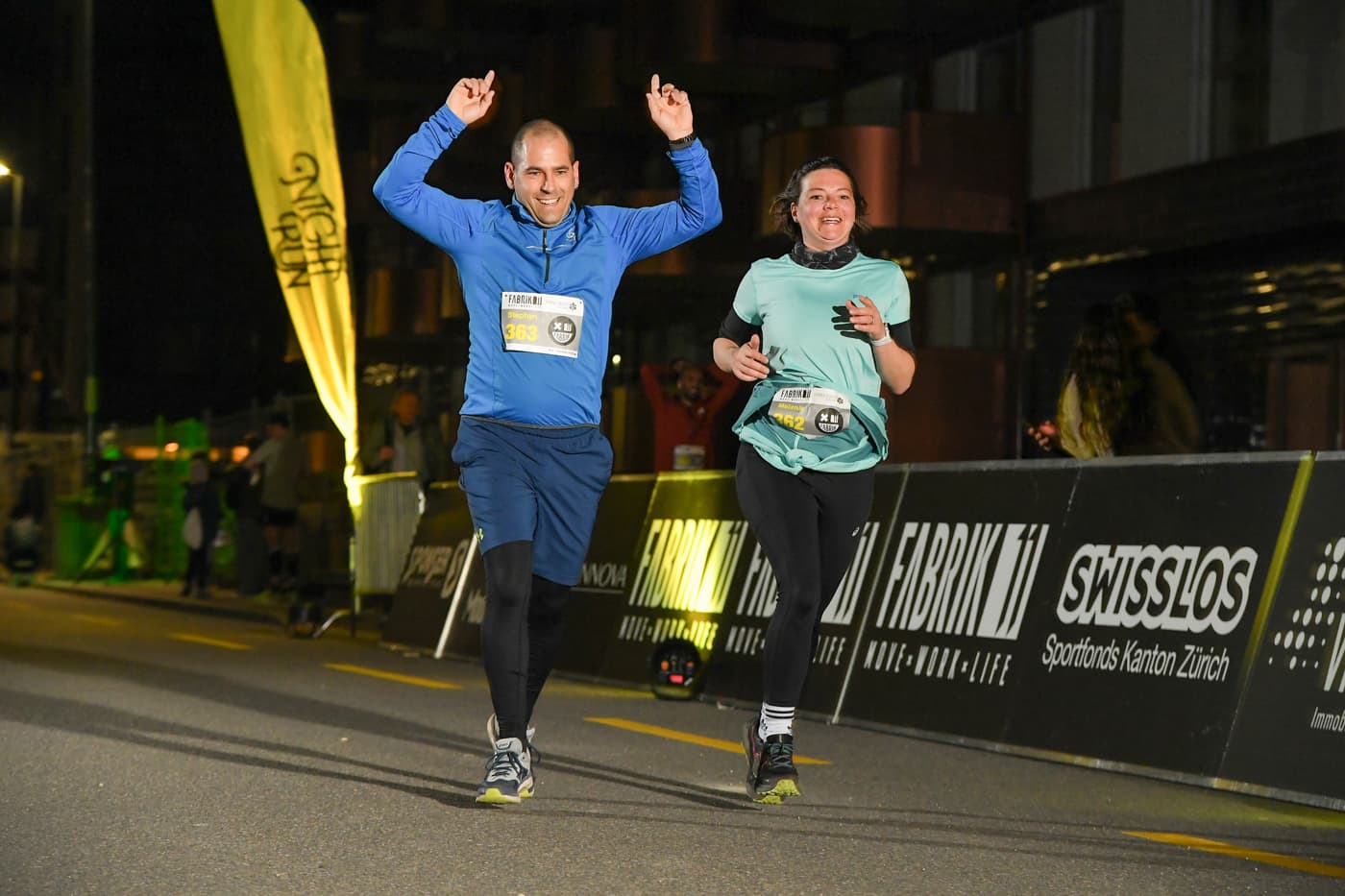 Zwei Läufer jubeln im Ziel des NightRun Zürich 2025 vor den Sponsorenbanden in Dübendorf