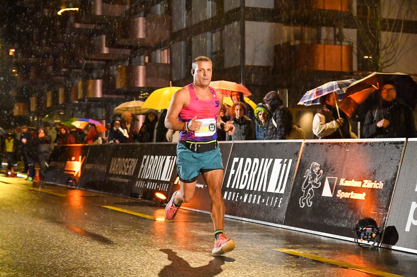 Läufer in Pink rennt im Regen an Zuschauern mit Schirmen vorbei, NightRun Zürich in Dübendorf