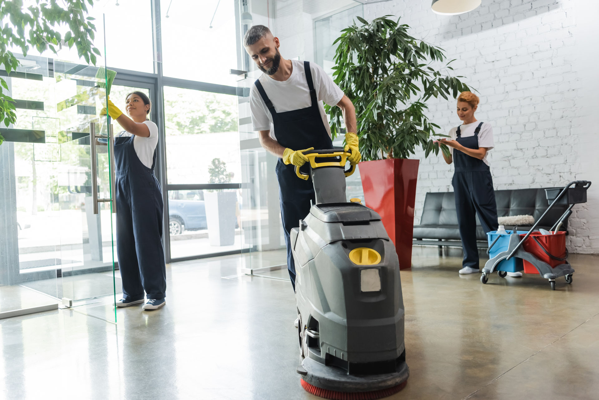 Cleaning team using a floor scrubber and wiping glass in a bright office foyer