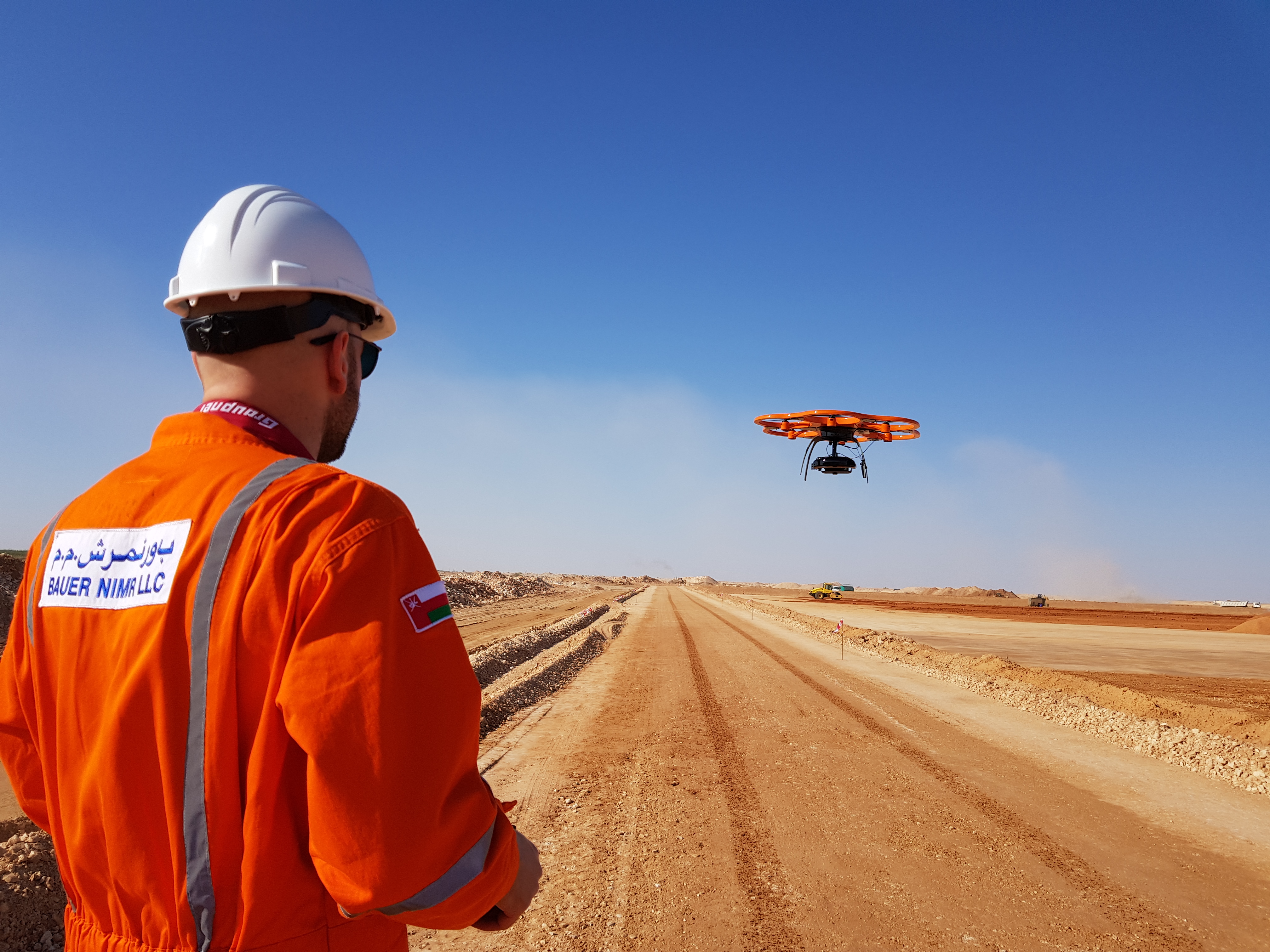 BAUER NIMR team member in orange overalls and a white helmet watching a survey drone fly over a site in Oman