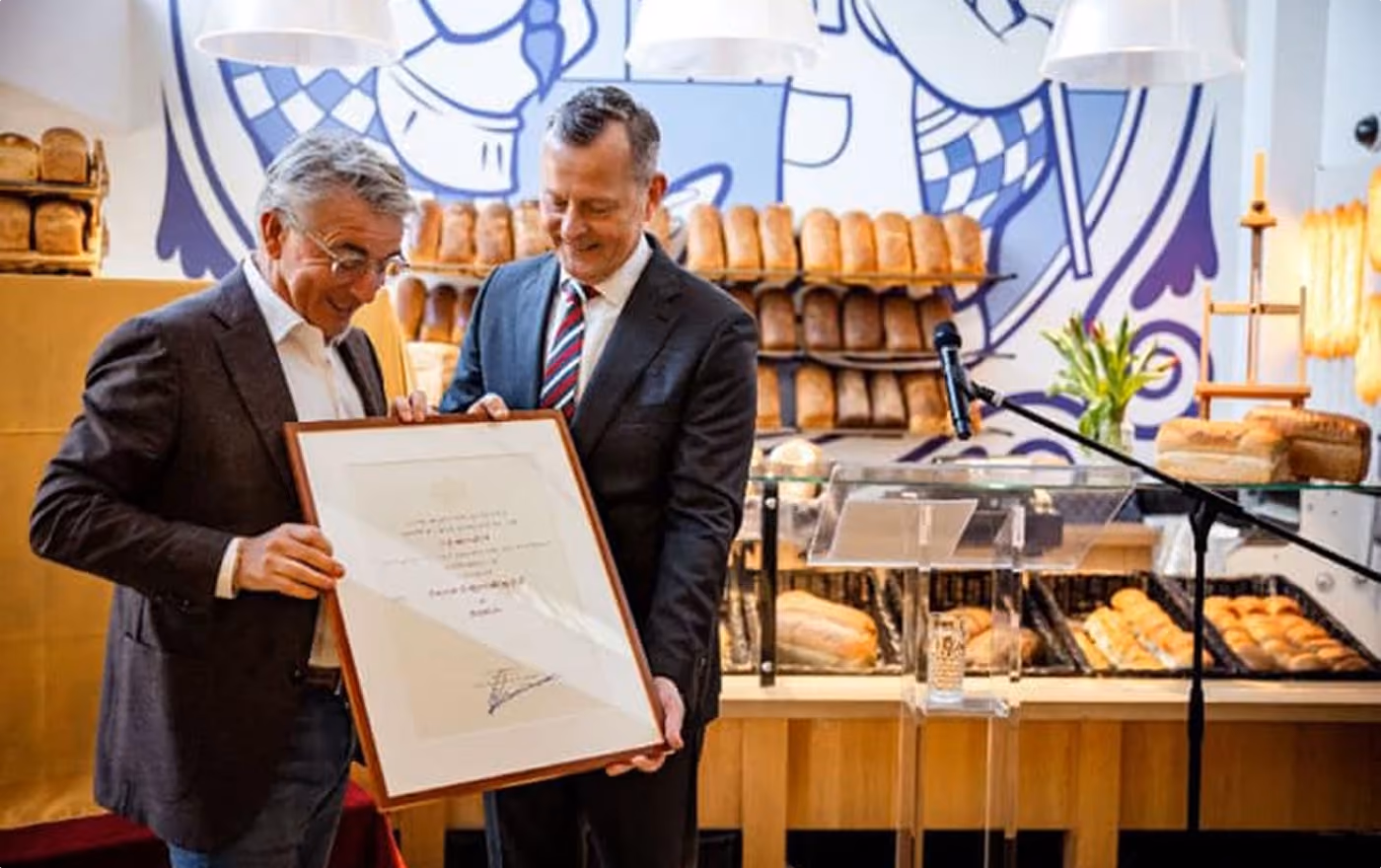 Two men in a bakery holding and looking at a large framed certificate; shelves of bread and a microphone stand are in the background.