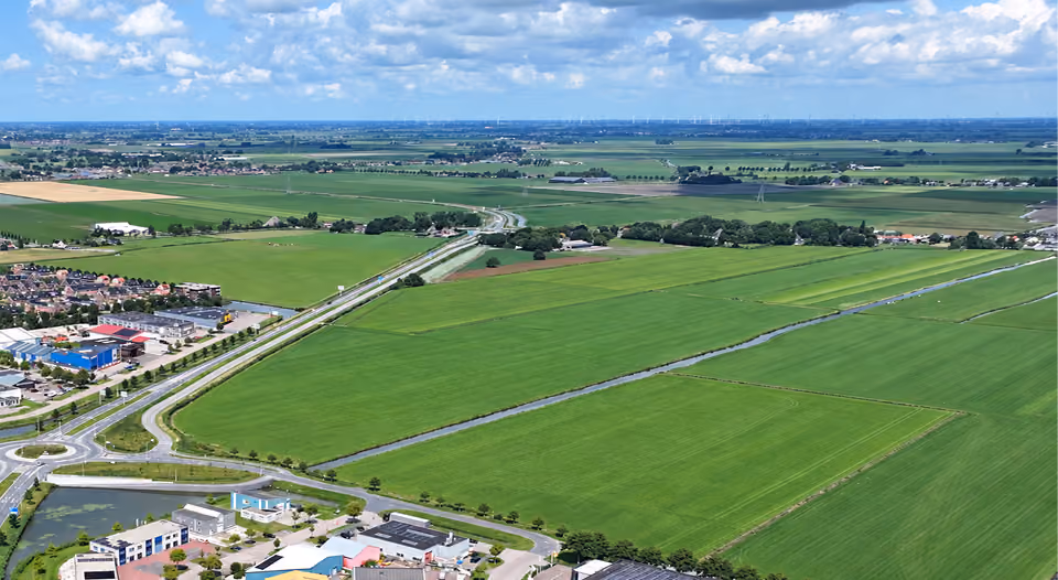 Aerial view of expansive green agricultural fields intersected by narrow canals with a cluster of buildings and roads in the foreground, under a blue sky with scattered clouds.