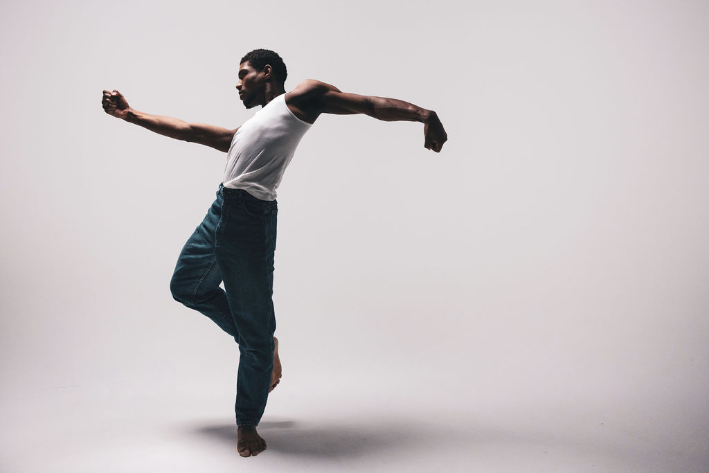 A dancer balanced on one leg with both arms extended, in a white tee and blue jeans, from the BNFTS studio shoot