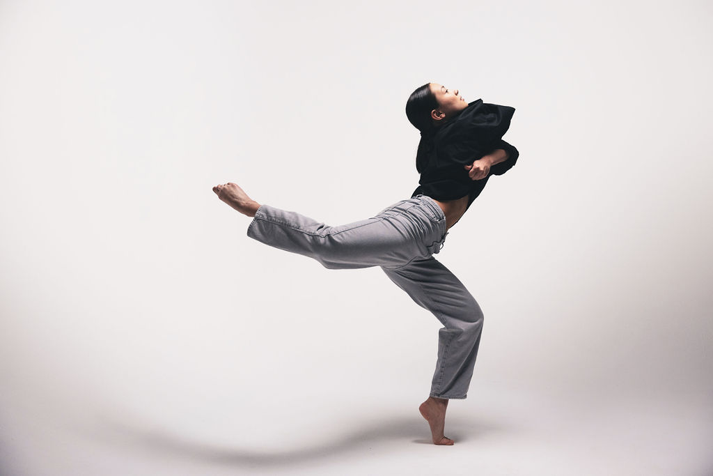 A dancer mid-kick with one leg extended, in a black hooded top and grey trousers, from the BNFTS studio shoot
