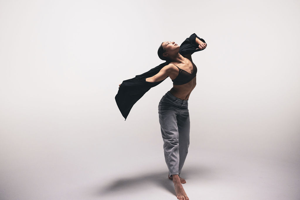 A dancer arching backwards in a black top and grey jeans, dark fabric trailing from one hand, from the BNFTS studio shoot