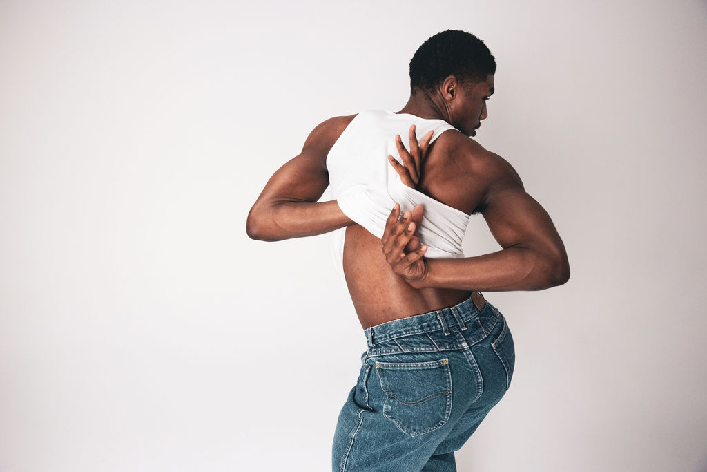 A dancer turned away, reaching behind their back, in a white tank and blue jeans, from the BNFTS studio shoot