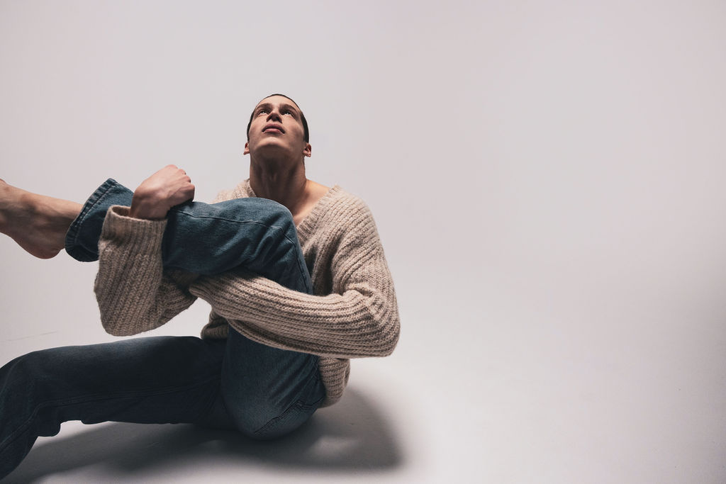 A dancer seated on the floor in a knit sweater, looking upward, from the BNFTS studio shoot