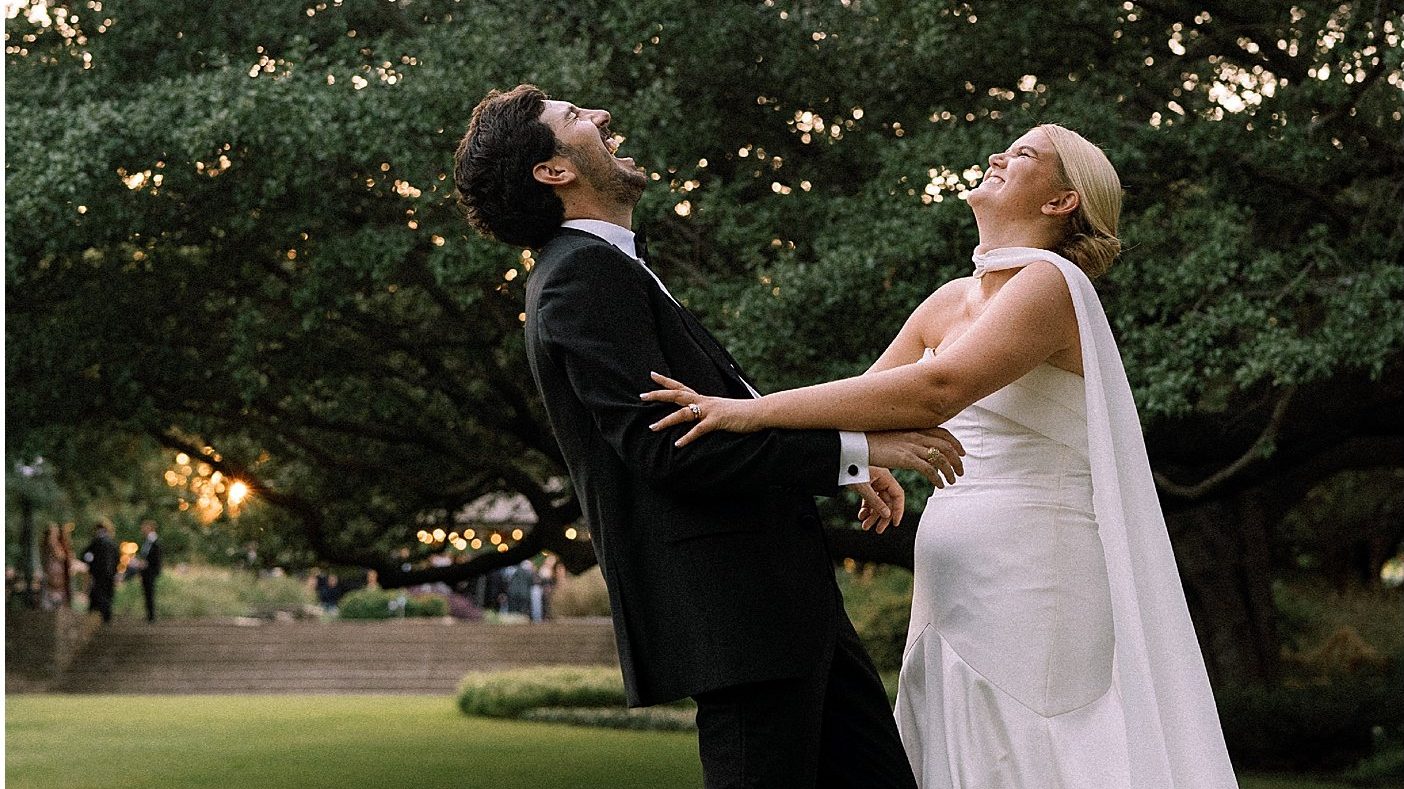 A couple laughing under live oaks strung with lights.