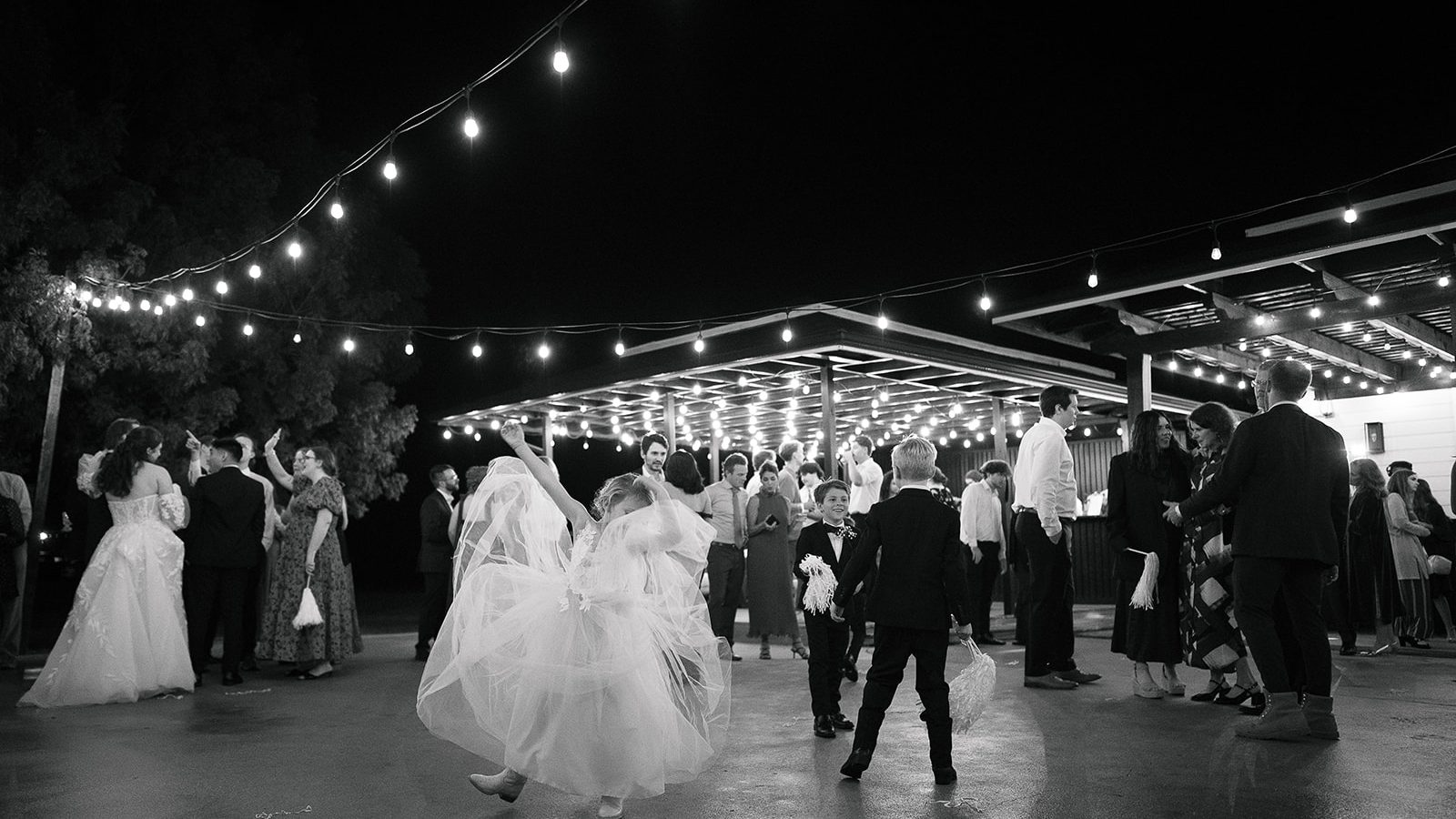 Black and white frame of the couple dancing on a plaza under string lights, with guests around them