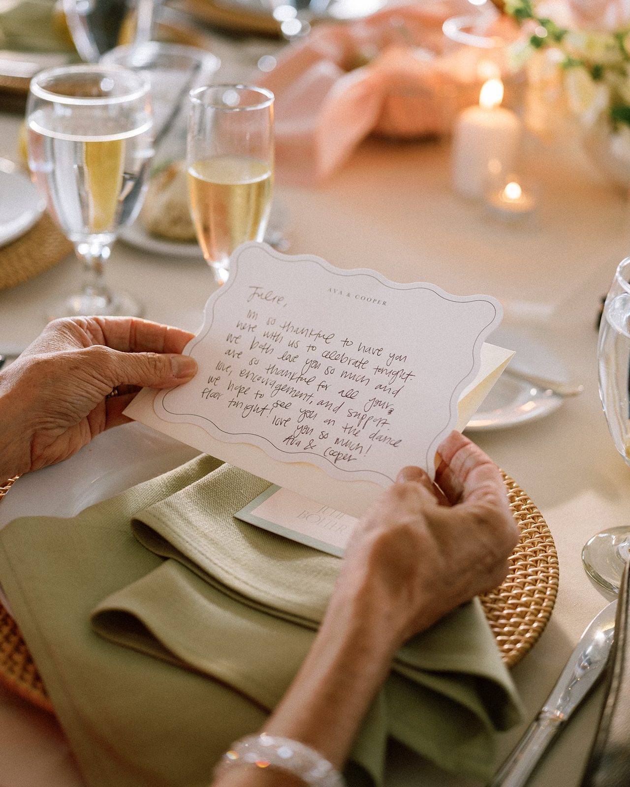 Hands holding a handwritten note at a wedding table, a green napkin and champagne glasses beside it