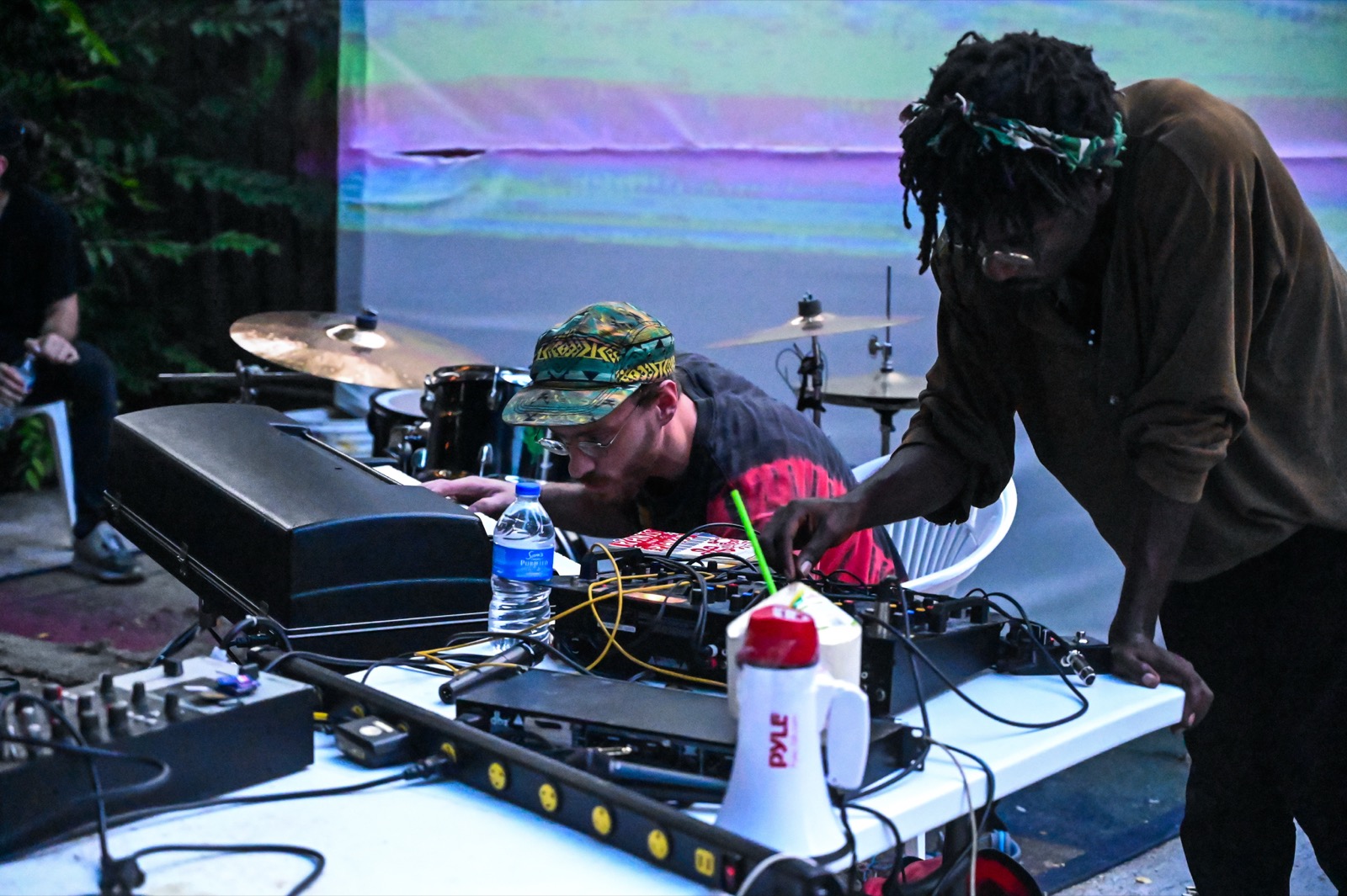 Musicians working a table of gear at a DIY show, a drum kit behind them