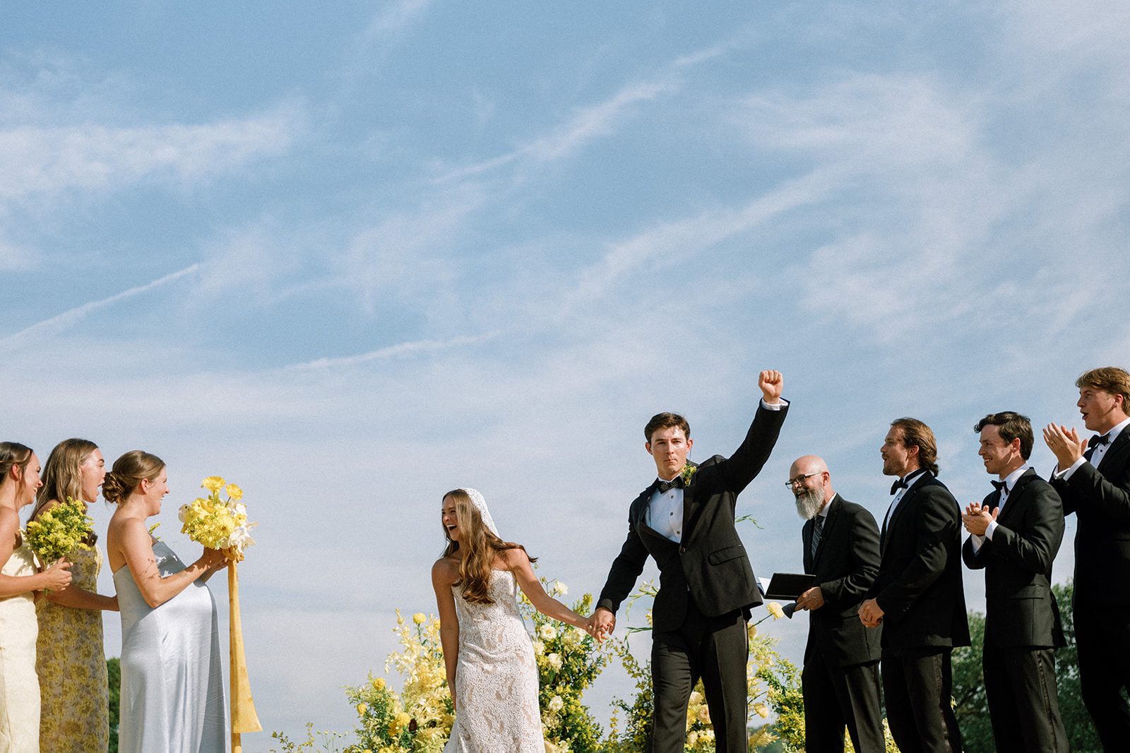 The groom raises a fist as the bridal party gathers on a hilltop under a wide blue sky