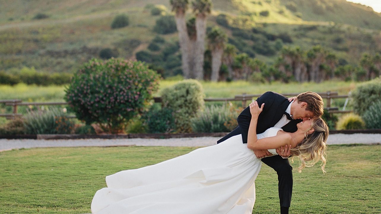 The groom dips the bride on a green lawn with palms and hills behind