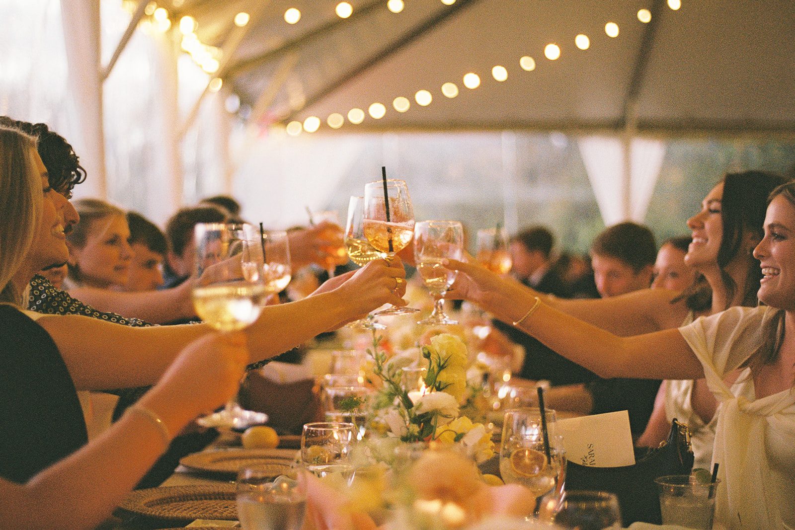 Guests reach across a long table to toast with glasses beneath a tent strung with lights
