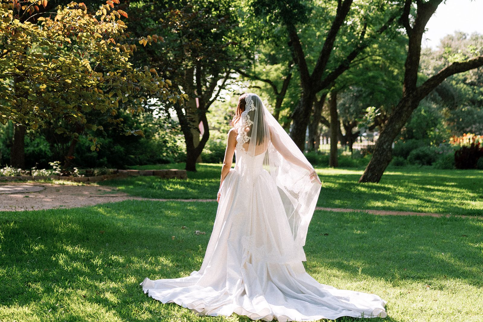 The bride from behind in a long veil, walking across a green lawn beneath tall trees