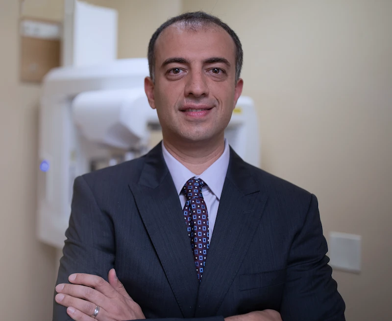 Dr. Ilya Ames, DDS, owner of Queens Central Dental in Jackson Heights, standing in a suit with arms crossed in his office