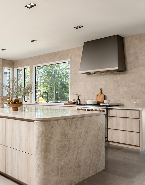 Modern kitchen with beige stone island, stainless steel range hood, and large windows showing greenery outside.