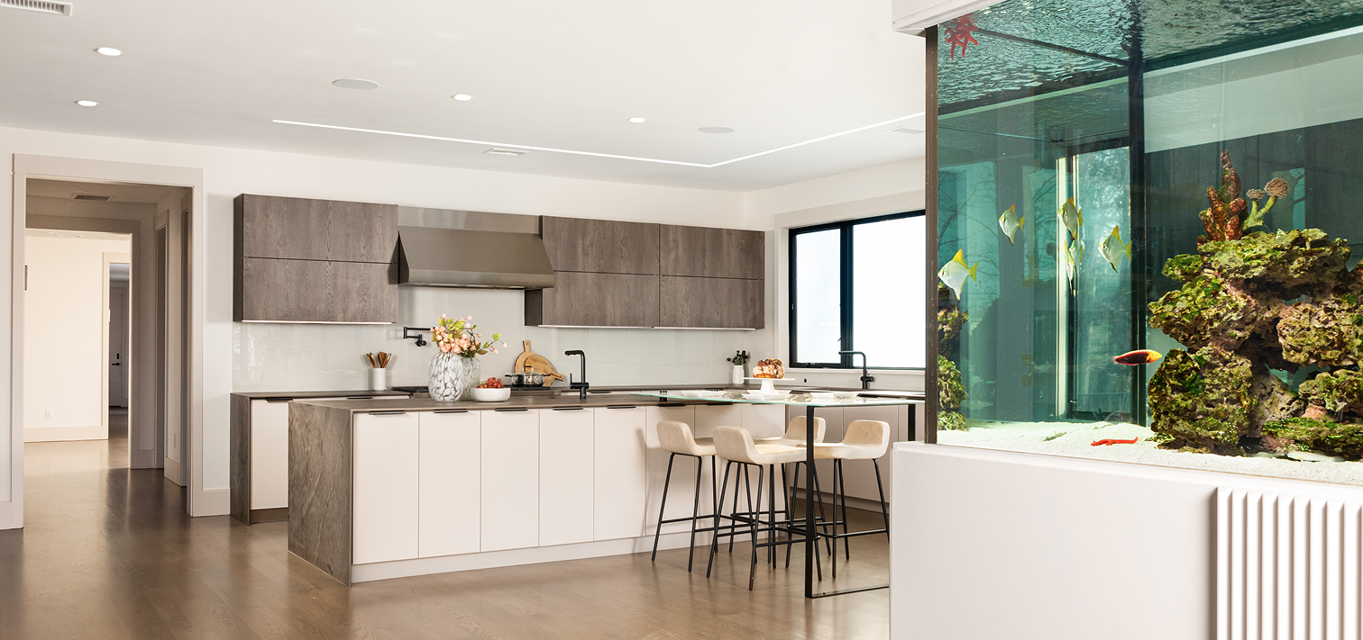 Modern kitchen with light wood floors, an island with stools, dark upper cabinets, and a large built-in aquarium with fish.