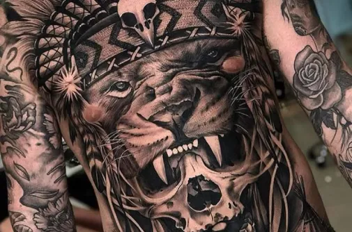 Torso tattoo of a roaring lion in a feathered headdress above a skull