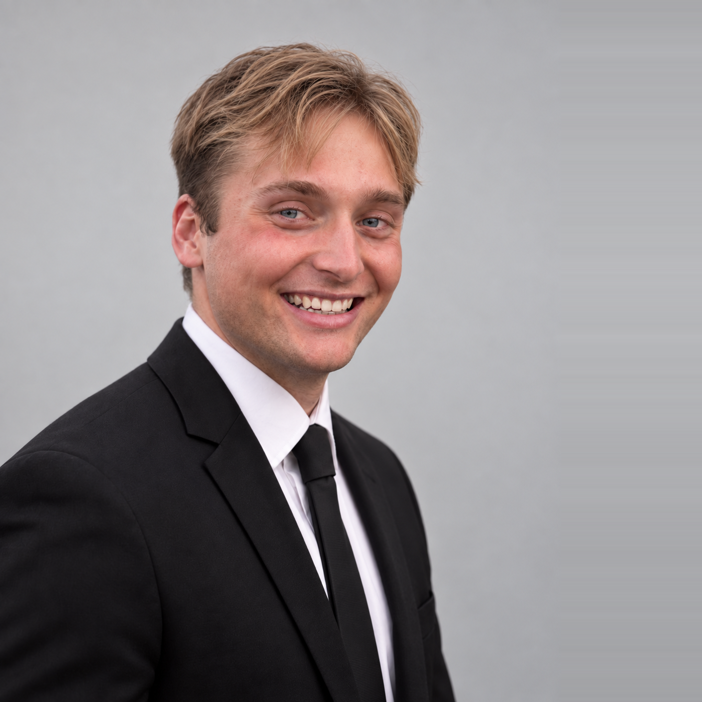 Young man with light brown hair wearing a black suit, white shirt, and black tie smiling against a gray background.