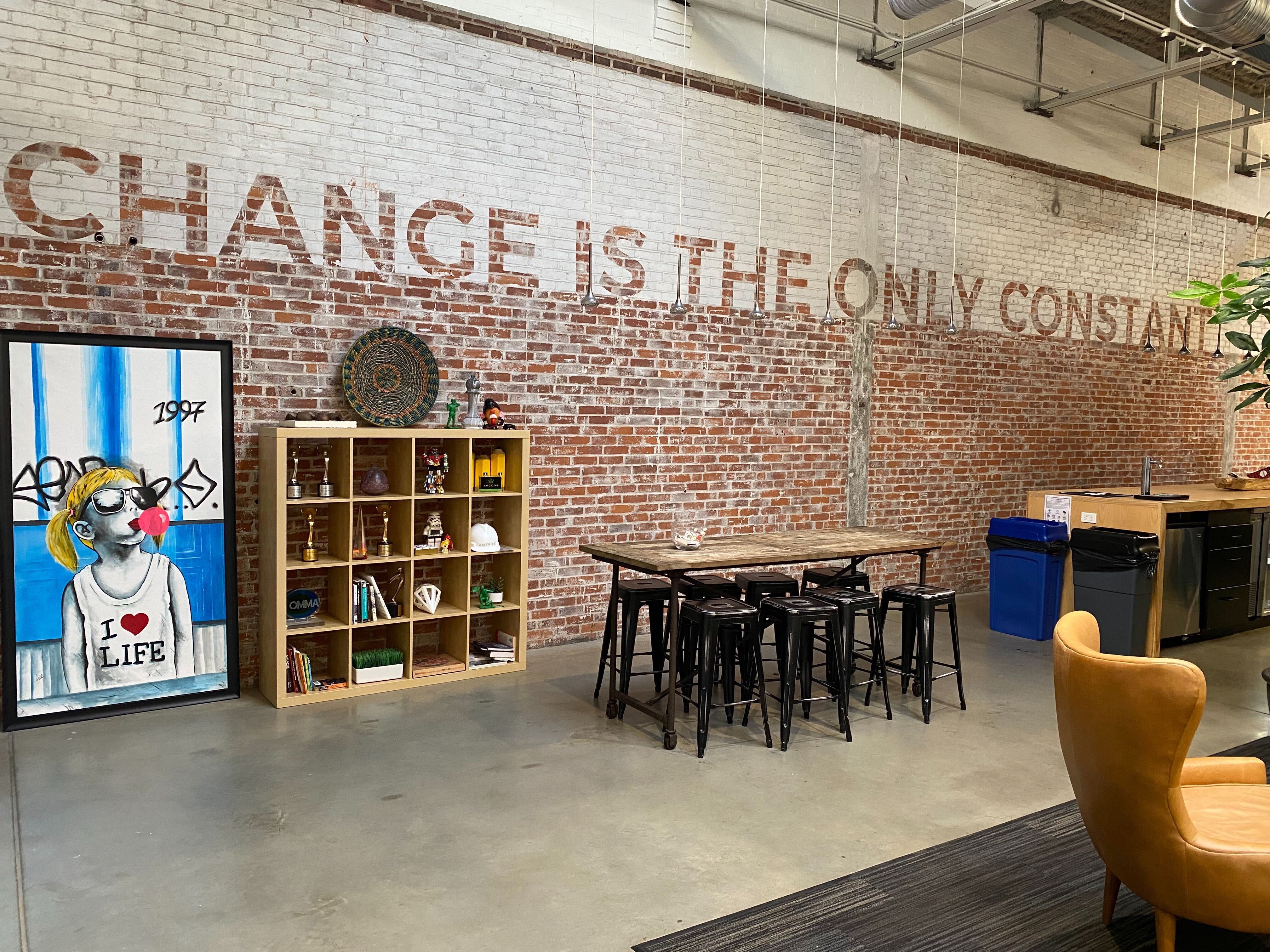 Picture of a kitchen space showing a brick wall with the words "Change is the only Constant" sand blasted into the red and white bricks. Leaning against the wall is a framed drawing of a girl in black and white chewing bubble gum wearing sunglasses, and next to the framed drawing of the girl is a bookshelf with trophies on each shelf.