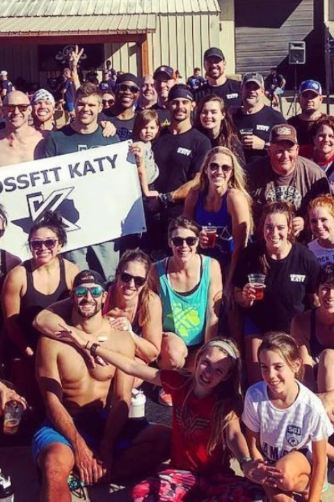 Large group of people posing outdoors in front of a building, many wearing CrossFit Katy apparel, with some holding a CrossFit Katy banner, smiling and enjoying a sunny day.