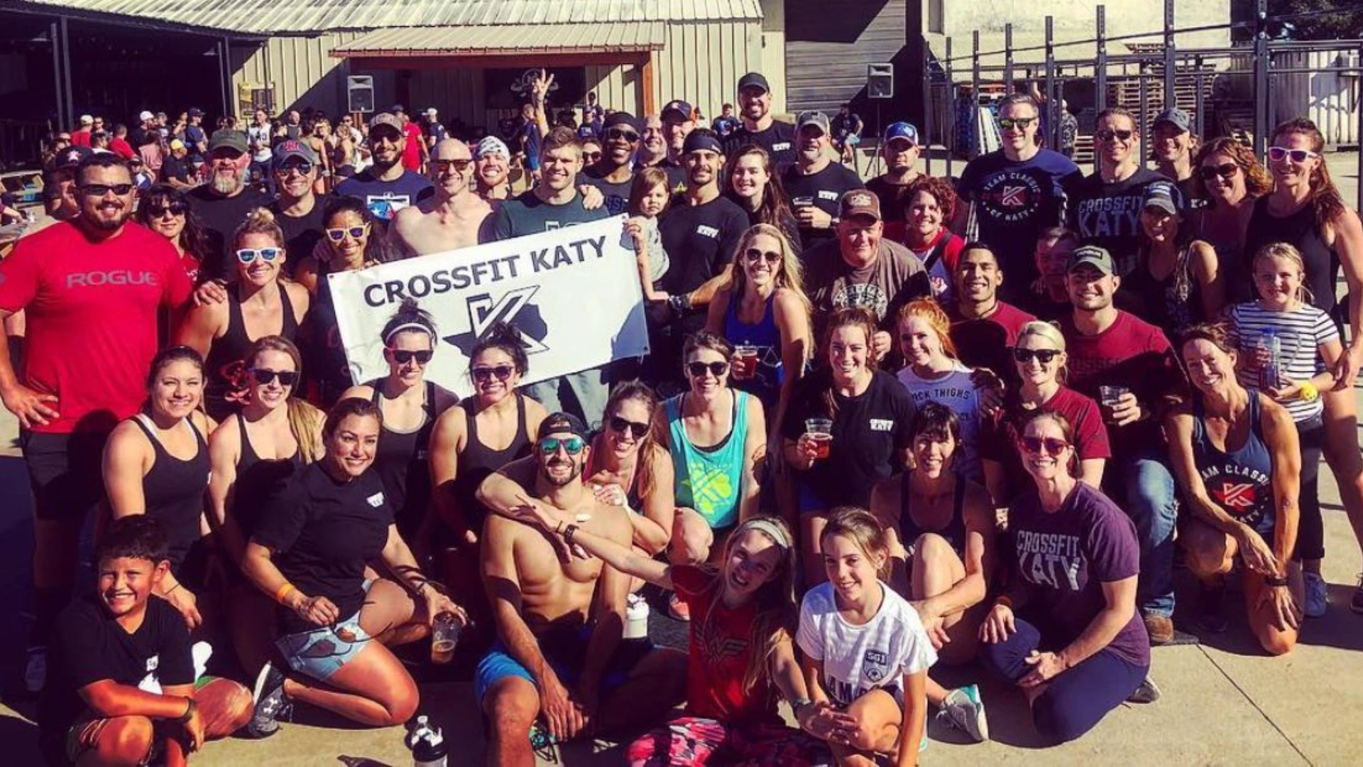Large group of people posing outdoors in front of a building, many wearing CrossFit Katy apparel, with some holding a CrossFit Katy banner, smiling and enjoying a sunny day.