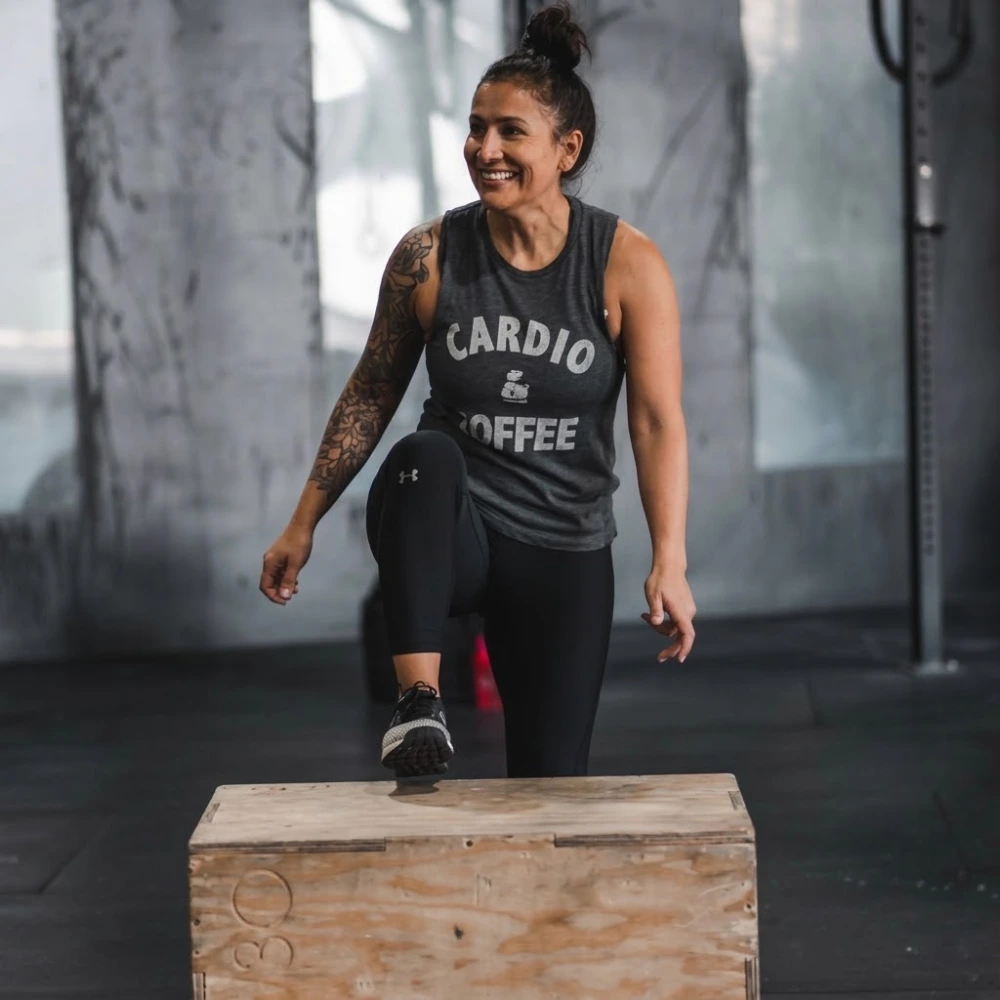 Smiling woman with a tattoo on her arm, wearing a sleeveless shirt with 'CARDIO & COFFEE' text, stepping onto a wooden plyometric box in a gym.