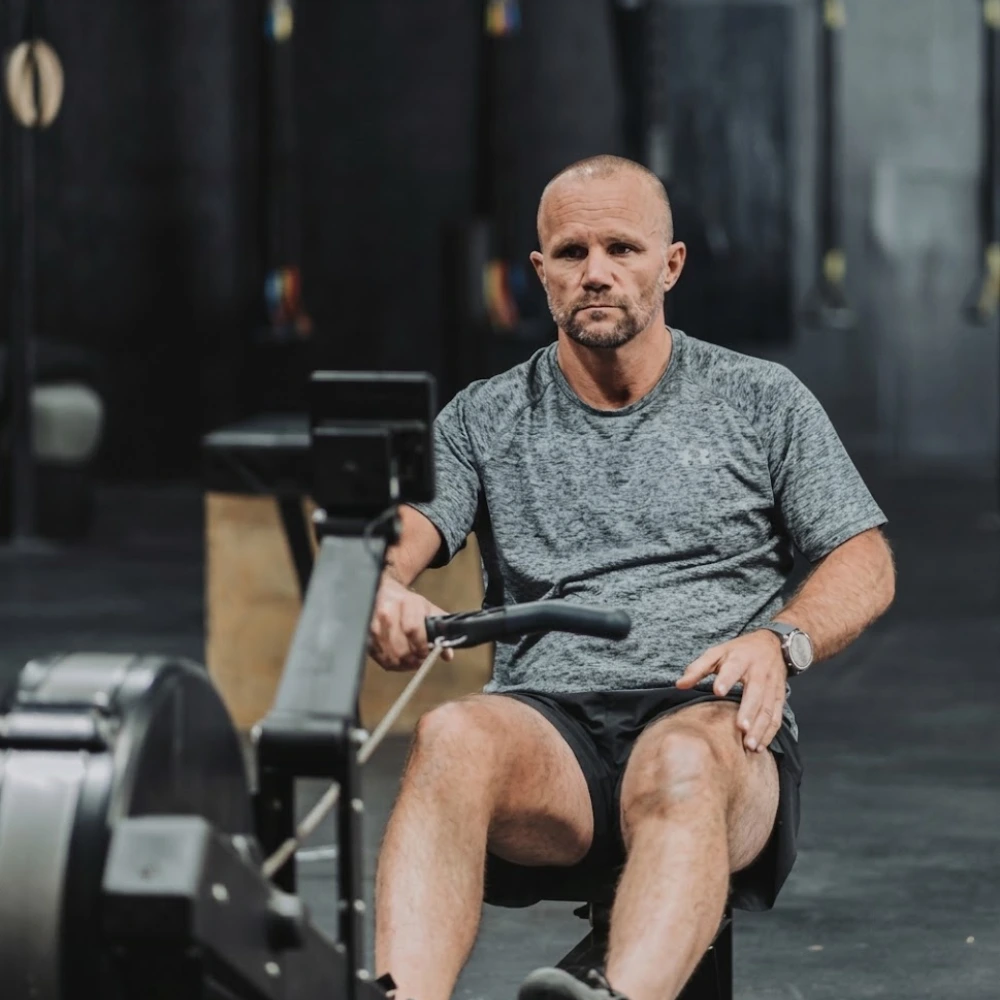 Athletic man with a shaved head and beard using a rowing machine in a gym setting, wearing a gray short-sleeve shirt and black shorts.