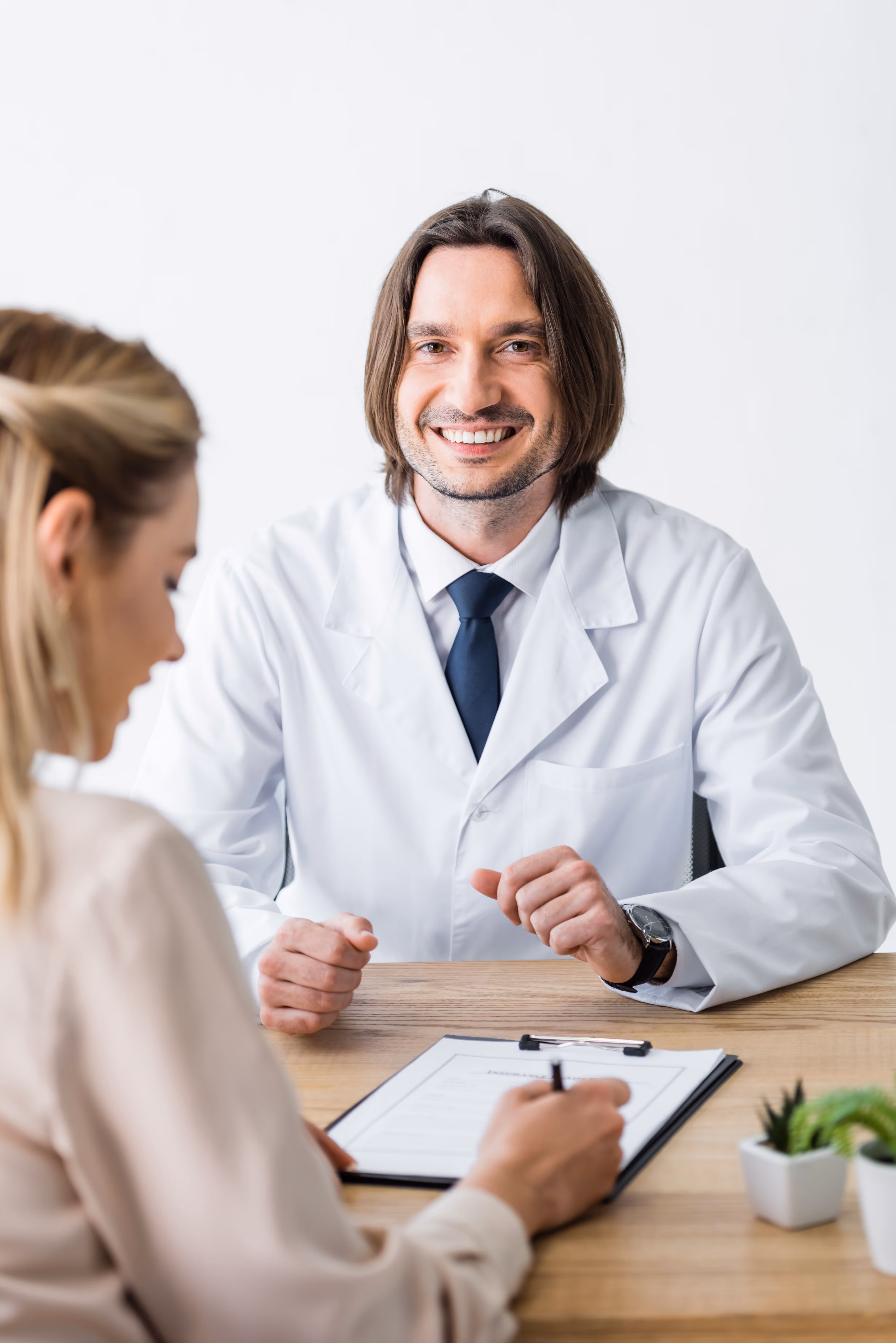 Smiling male doctor talking to a woman holding a pen and clipboard in a bright office.